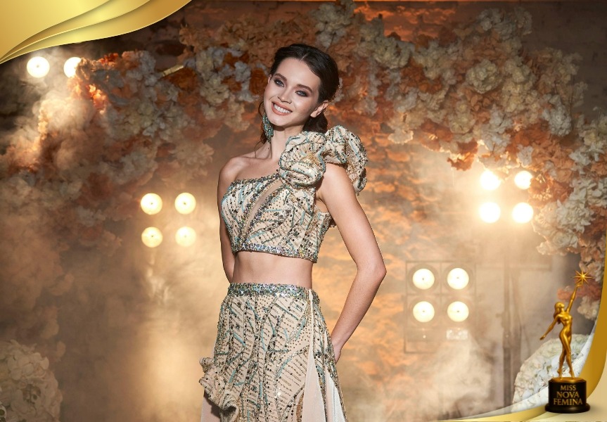 Concursante de Miss Nova Femina luciendo un conjunto elegante y bordado, en un escenario iluminado con detalles florales.